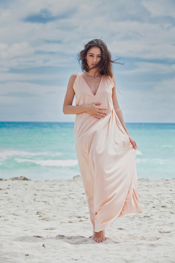 Eine Frau geht an einem malerischen Strand bei warmem Sommerwetter. Sie trägt ein langes, luftiges Sommerkleid, das sanft vom Wind umspielt wird. Ihre Haare wehen ebenfalls im Wind