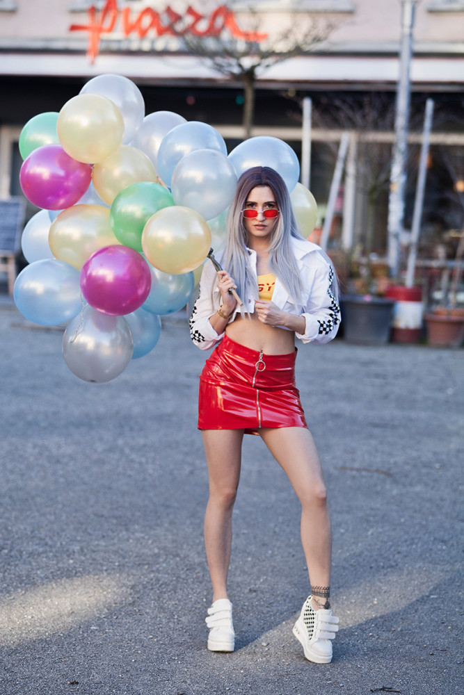 Eine junge Frau bei einem Streetfotoshooting an einem sonnigen Sommertag. Sie trägt einen auffälligen roten Minirock, kombiniert mit einem sommerlichen Oberteil. In der Hand hält sie eine Vielzahl bunter Luftballons